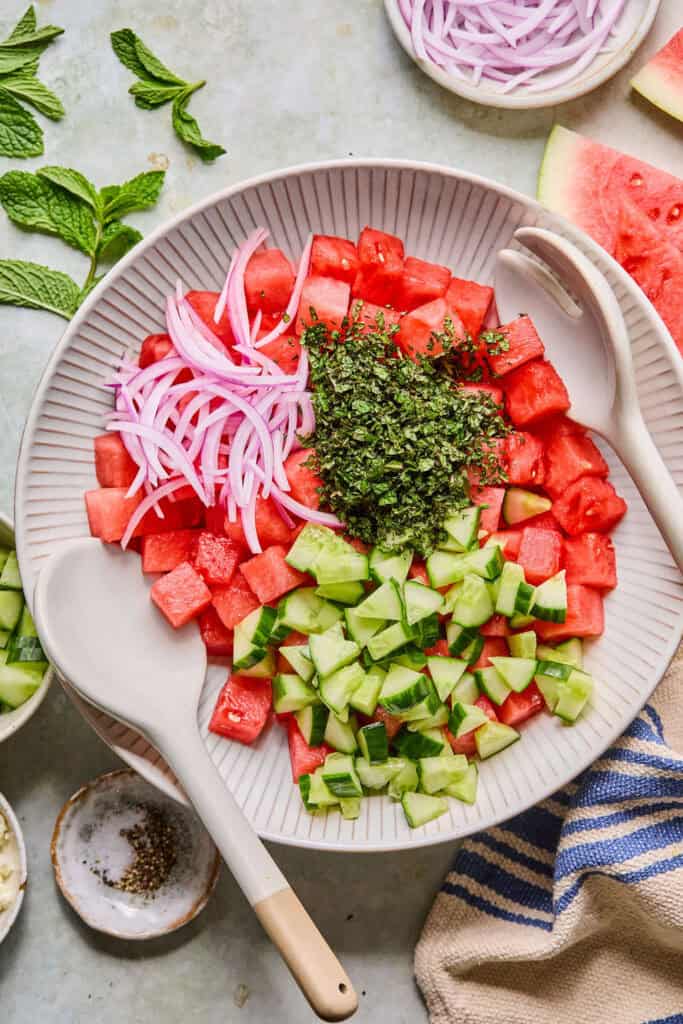 Watermelon cubes, cucumber, and red onion in a large bowl before being mixed together.