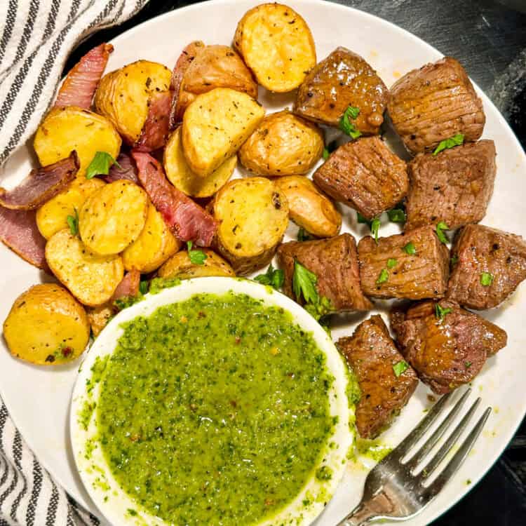Chimichurri Steak Bites with red potatoes and a side of chimichurri sauce on a plate with a fork.