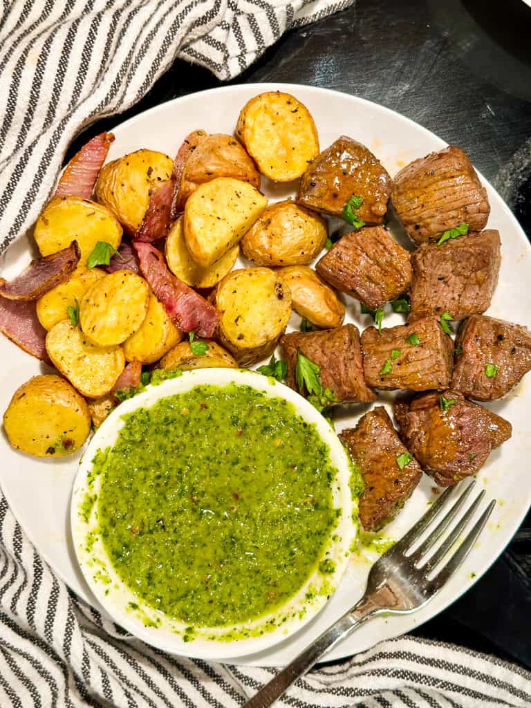 Chimichurri Steak Bites with red potatoes and a side of chimichurri sauce on a plate with a fork.