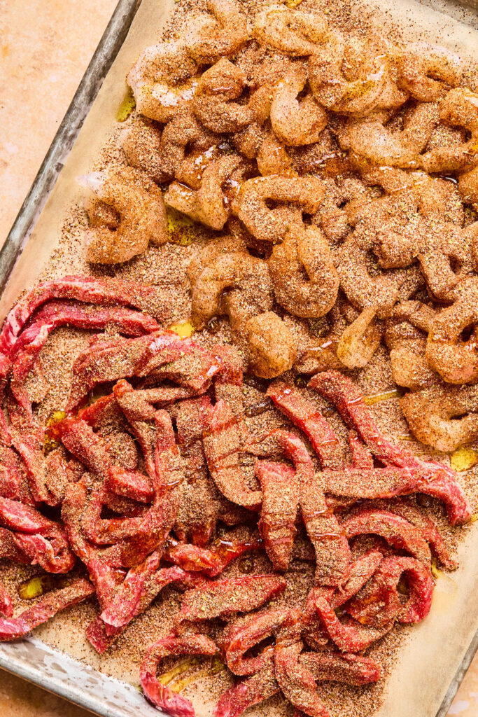 Raw steak and shrimp covered with spices and oil on a sheet pan with parchment paper.