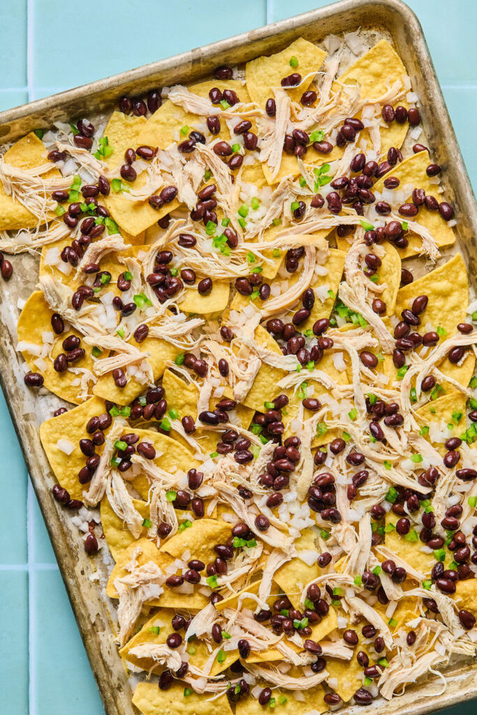 Tortilla chips on a baking sheet topped with shredded chicken, beans, and onion.