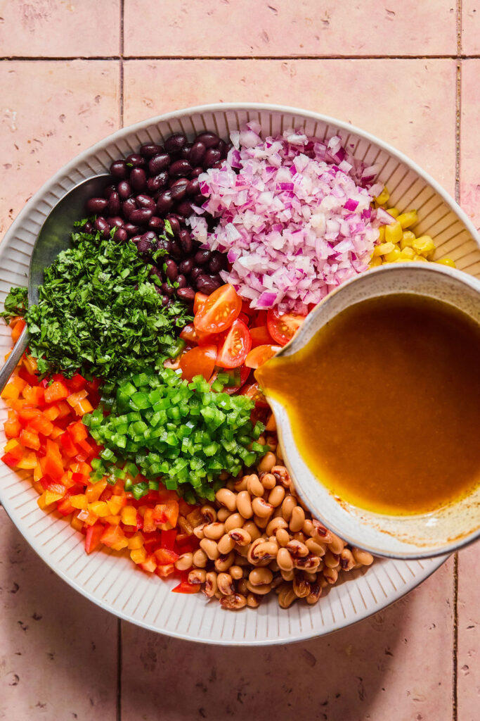 Dressing being poured over the Rainbow Cowboy Caviar ingredients in a large bowl.