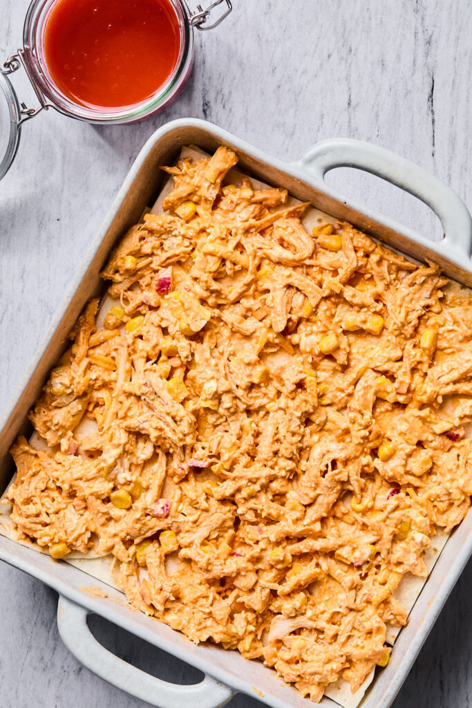 Chicken mixture on top of the tortillas in a square baking dish.